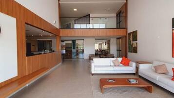 Ampla sala de estar com teto alto, sofá branco central, mesa de centro e vista para a cozinha.