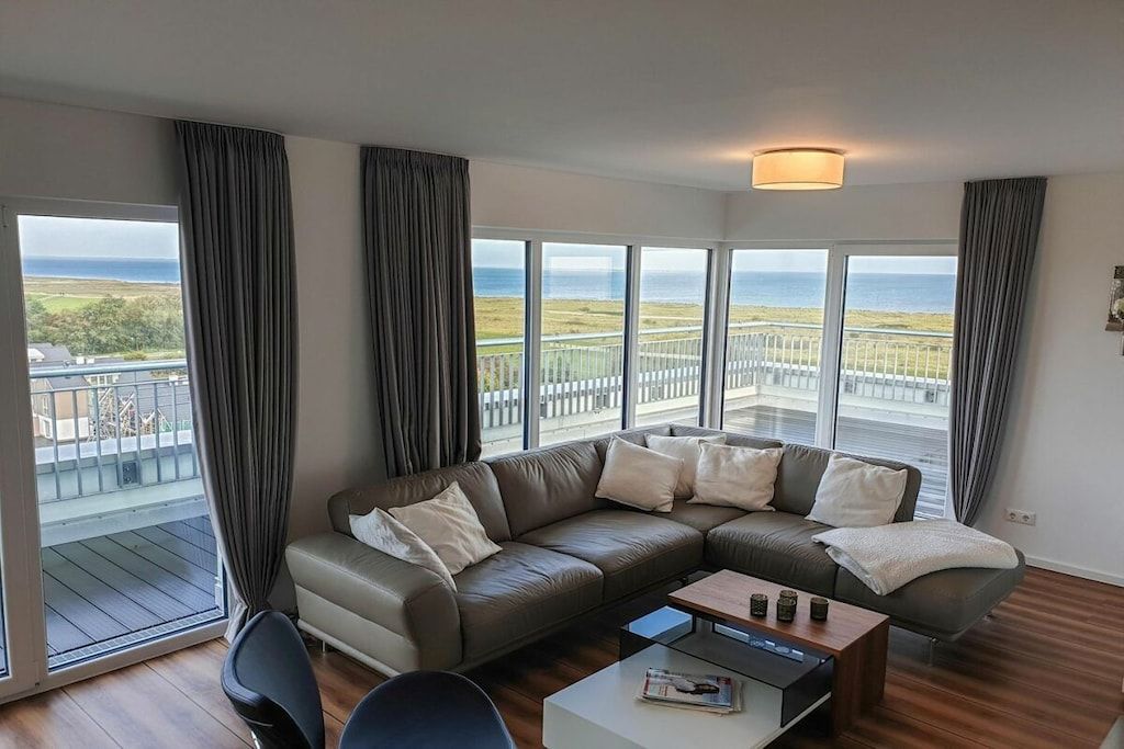 A modern living room with a large sectional sofa, a coffee table, and a view of the sea through large windows.