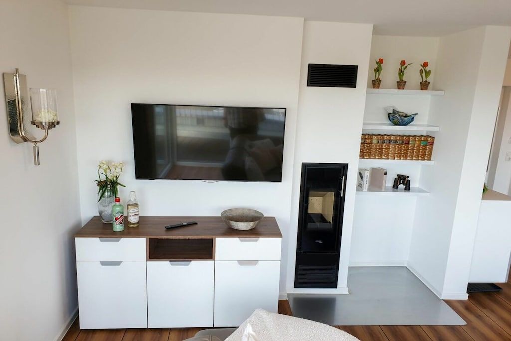 A modern living room with a flat-screen TV mounted on the wall, a wooden coffee table, a white cabinet with drawers, and a built-in fireplace.