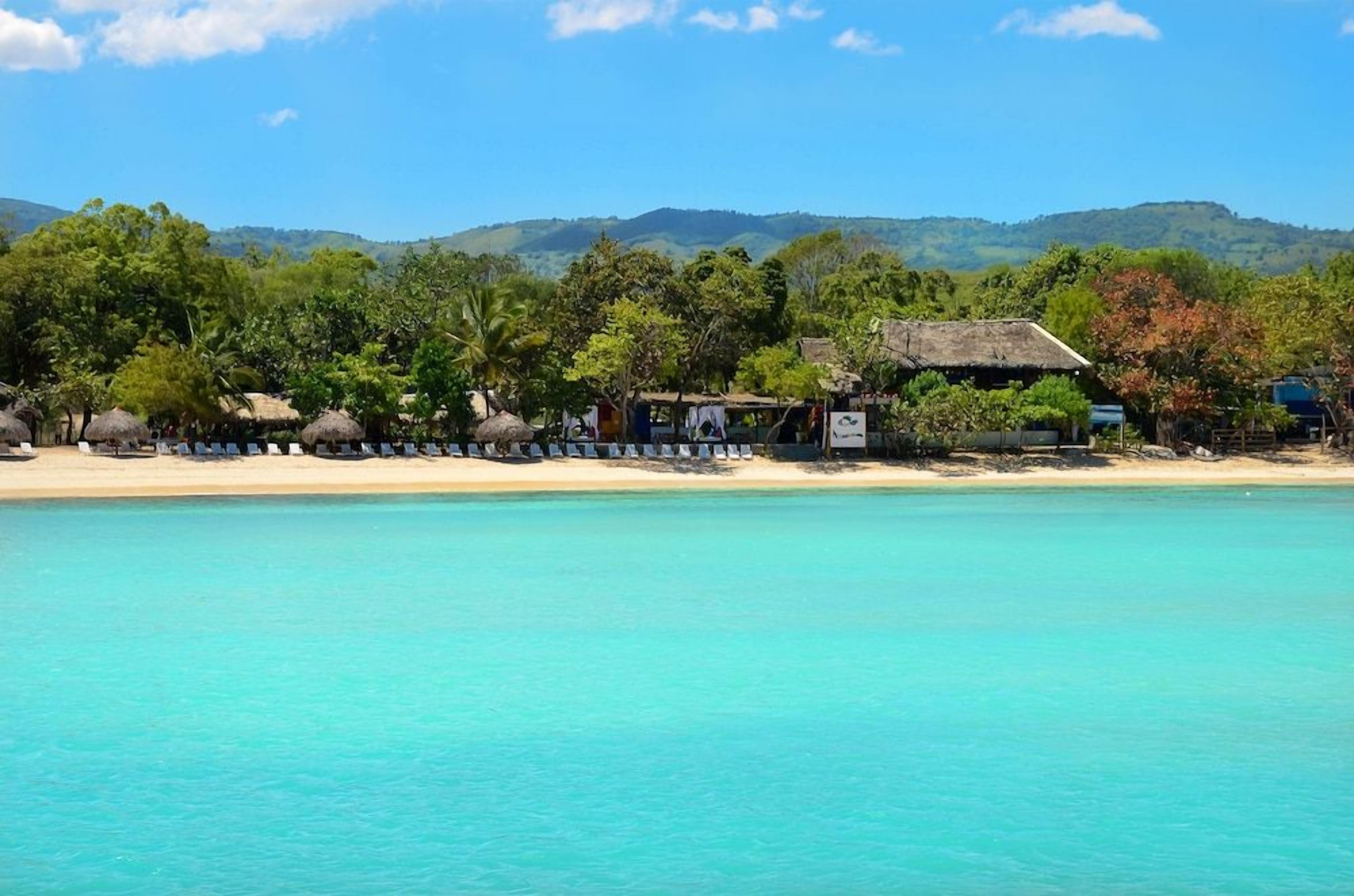 A beachside resort with thatched-roof huts, a sandy beach, and a backdrop of hills.