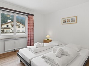 A bedroom with two beds, a window with a view of buildings, a radiator, and a bedside table with a lamp. - Seeli Appartement/Fewo,   Ferienwohnung Seeli IH by Interhome (Churwalden)