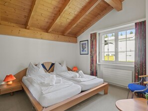 Una camera da letto con soffitto in legno, un letto con lenzuola bianche, un comodino con una lampada, una finestra con tende e una sedia.