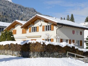 Ein zweistöckiges Chalet mit Natursteinfundament, Holzläden und einem mit Schnee bedeckten Dach.