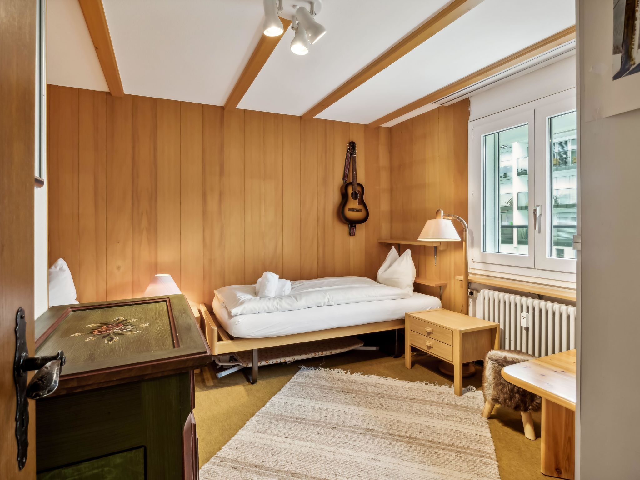 A bedroom with a wooden wall, a bed, a guitar, a lamp, a radiator, and a window.