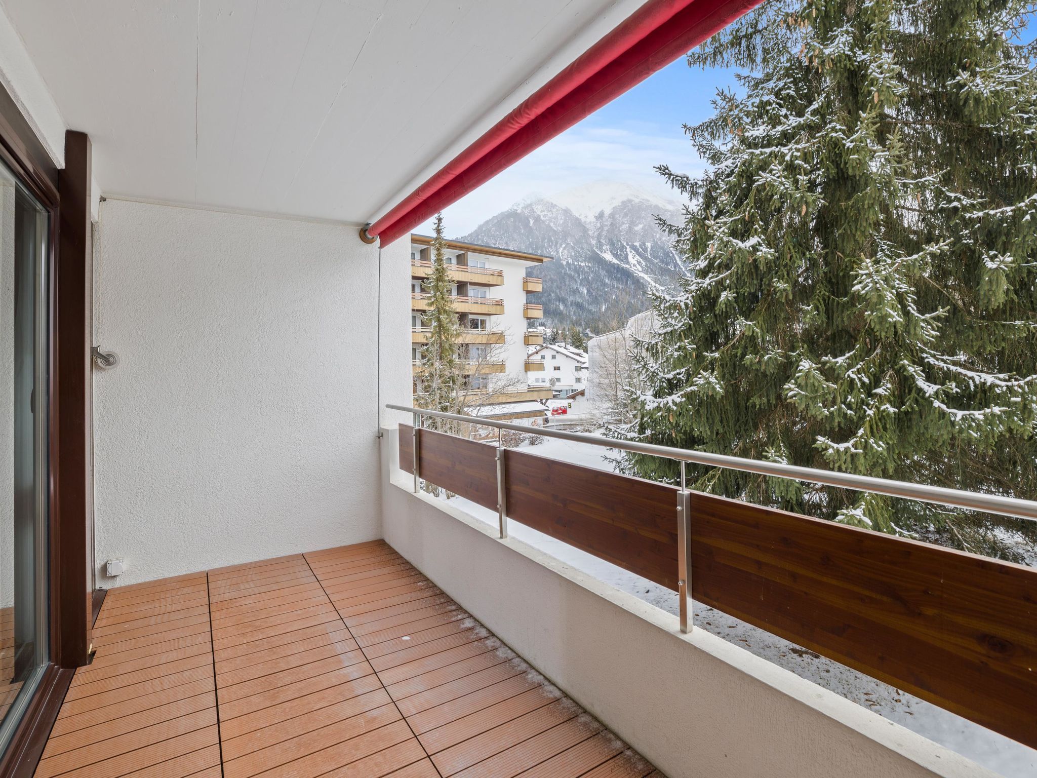A balcony with a wooden floor, a metal railing, and a view of snow-covered trees and mountains.