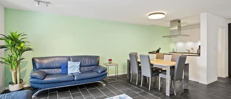 A modern living room with a sofa, dining table, chairs, and a kitchen area.