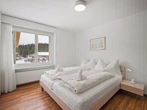 Ein Hotelzimmer mit einem großen Bett, einem Nachttisch mit Lampe, einem Fenster mit Blick auf Gebäude und Bäume sowie einem gerahmten Bild an der Wand.