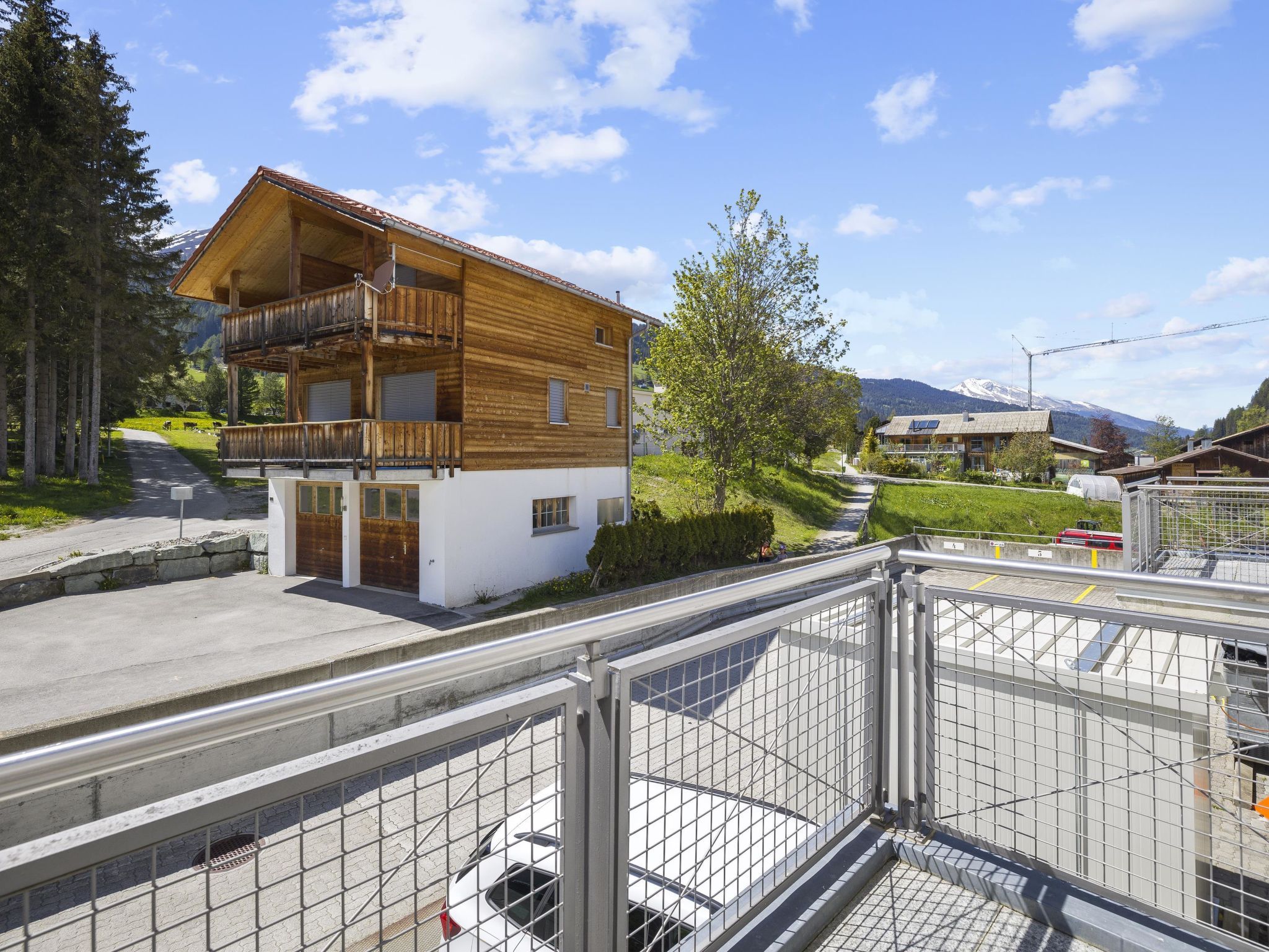 A two-story wooden chalet with a balcony, surrounded by greenery and a mountain backdrop.