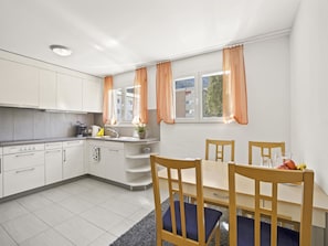 A modern kitchen with white cabinets, a dining table with chairs, and a window with orange curtains. - Moser Appartement/Fewo Ferienwohnung Moser IH by Interhome (Churwalden)