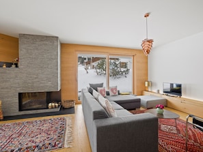 Ein modernes Wohnzimmer mit Kamin, einer großen Sofagarnitur, einem Fernseher und Blick auf eine verschneite Landschaft durch eine Glastür.