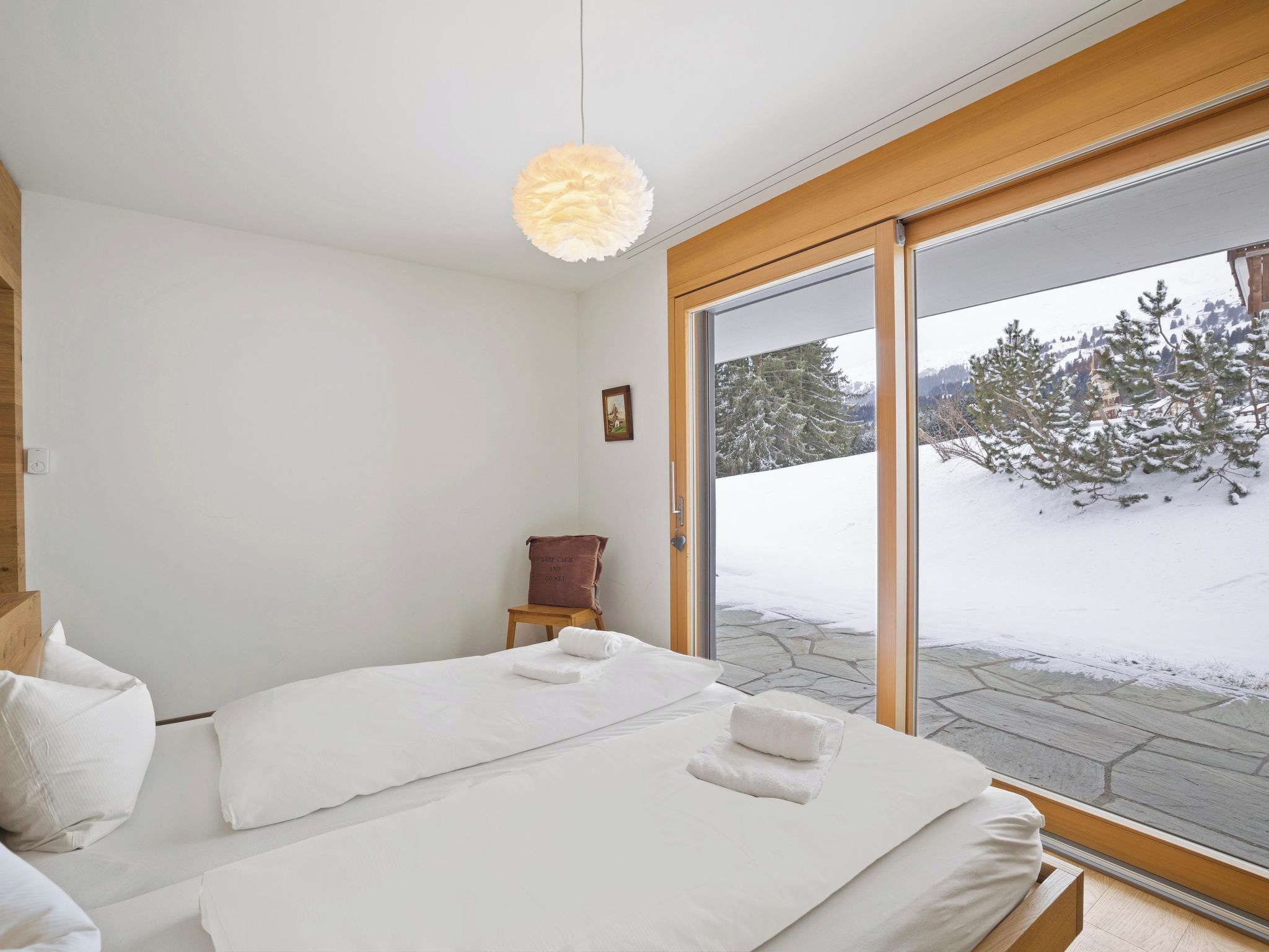 Een slaapkamer met een bed, een houten bankje en uitzicht op een besneeerd landschap door een groot raam.