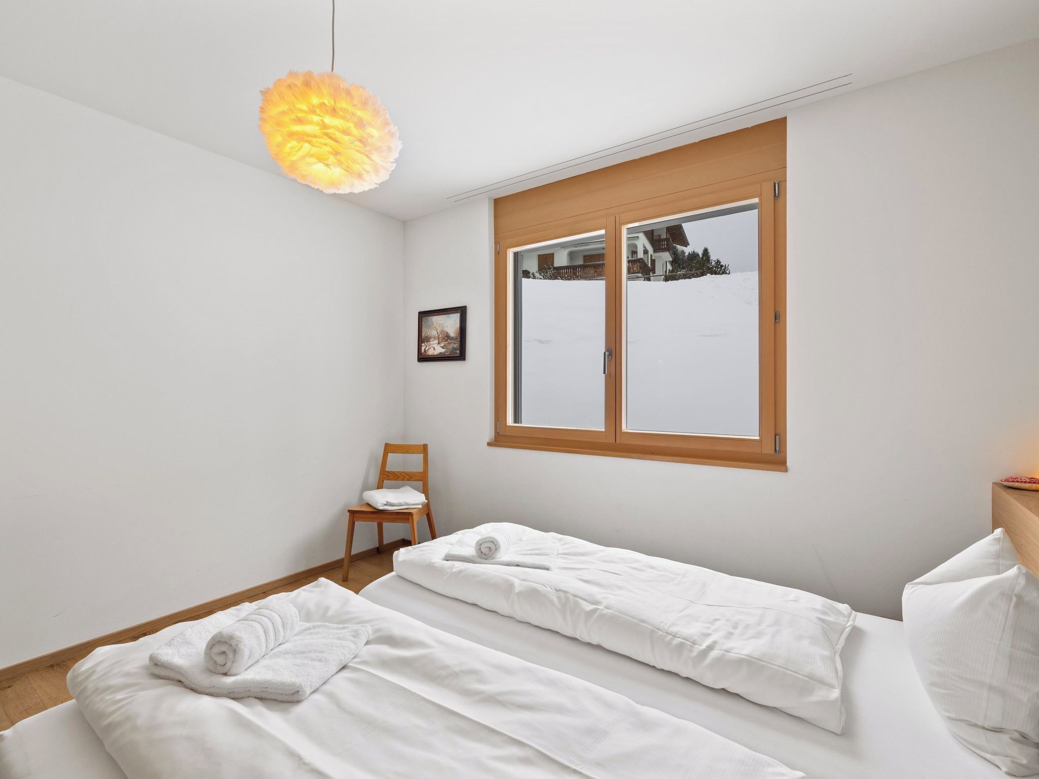 A bedroom with two beds, a chair, a painting, and a window with a view of a house and trees.