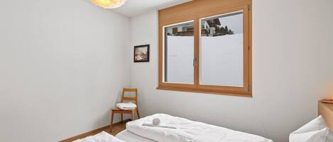 A bedroom with two beds, a chair, a painting, and a window with a view of a house and trees.