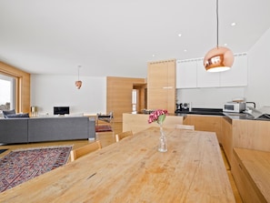 Eine moderne Küche mit Holzausstattung, ein Essbereich mit großem Holztisch und ein graues Sofa.