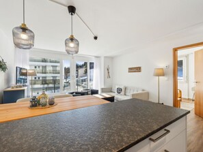 A modern kitchen with a large island, pendant lights, and a view of a residential building through large windows. - Nr.06 Haus Sursilvana Appartement/Fewo,   Nr.06 Haus Sursilvana IH by Interhome (Lenzerheide)