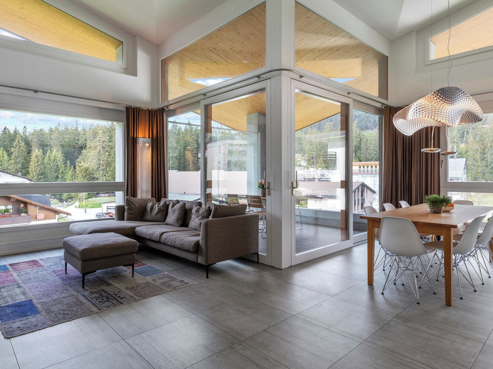 A modern living room with a sofa, a dining table with chairs, and a view of a forest through large windows.
