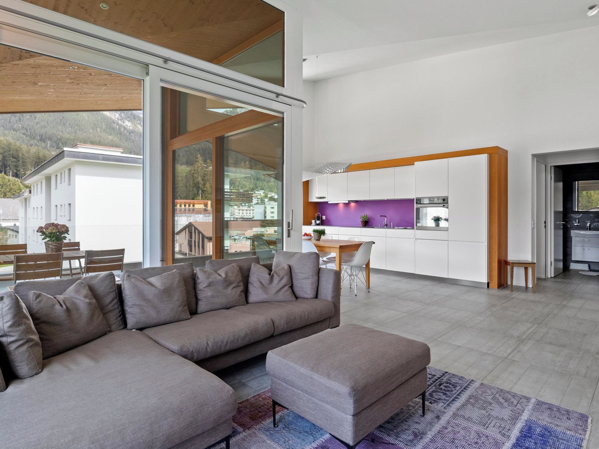 Un saló modern amb un sofà seccional i un pouf, una zona de menjador amb taula i cadira, i una cuina amb armaris blancs i una paret de color morat.