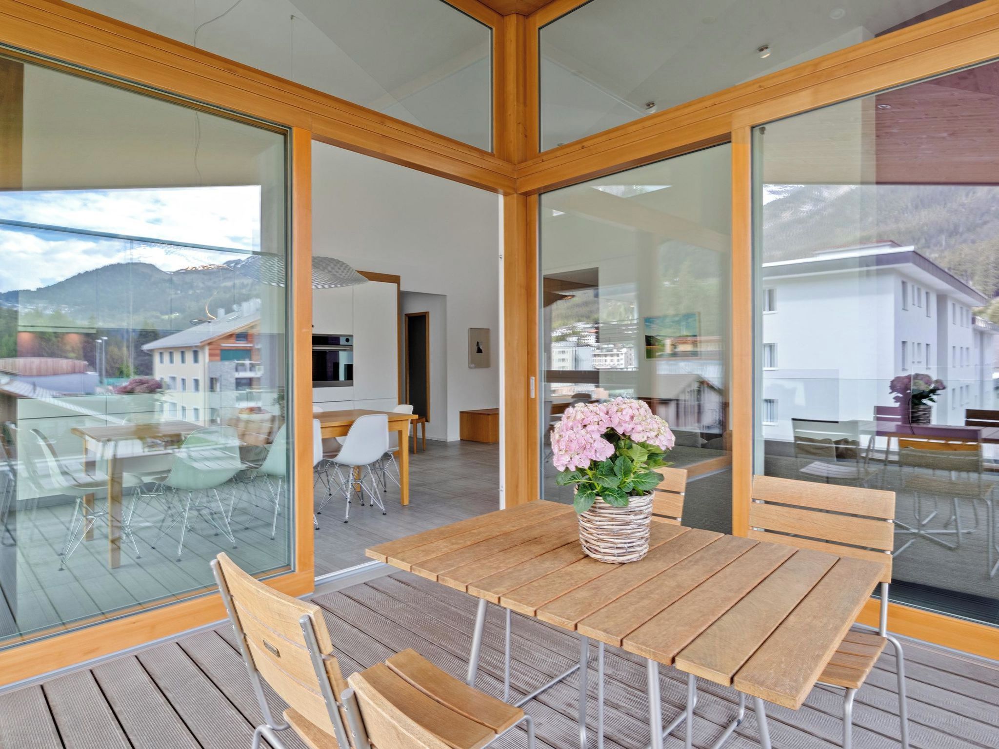 Een modern interieur met een houten tafel en stoelen, een bloemenvaas en grote glasramen met uitzicht op het omliggende landschap.