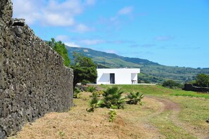 Um edifício moderno branco com porta preta, cercado por uma área gramada com algumas palmeiras, ao fundo colinas suaves e um céu claro.