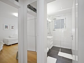 Ein modernes Badezimmer mit Duschkabine aus Glas, einem weißen Waschbecken und einer Toilette. Im angrenzenden Wohnbereich befindet sich eine weiße Couch.
