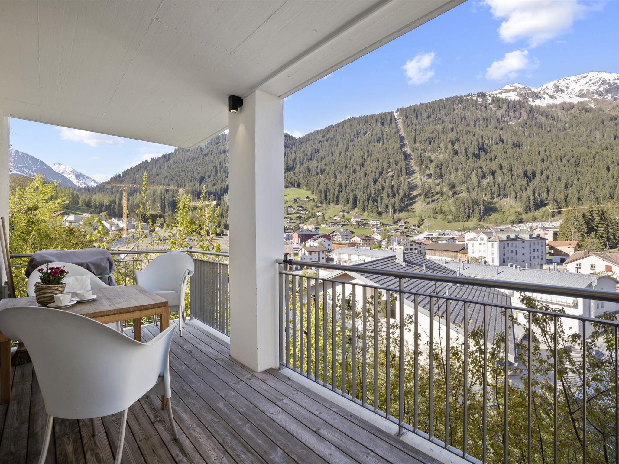 A balcony with a wooden floor, white chairs, and a table with a basket of flowers.