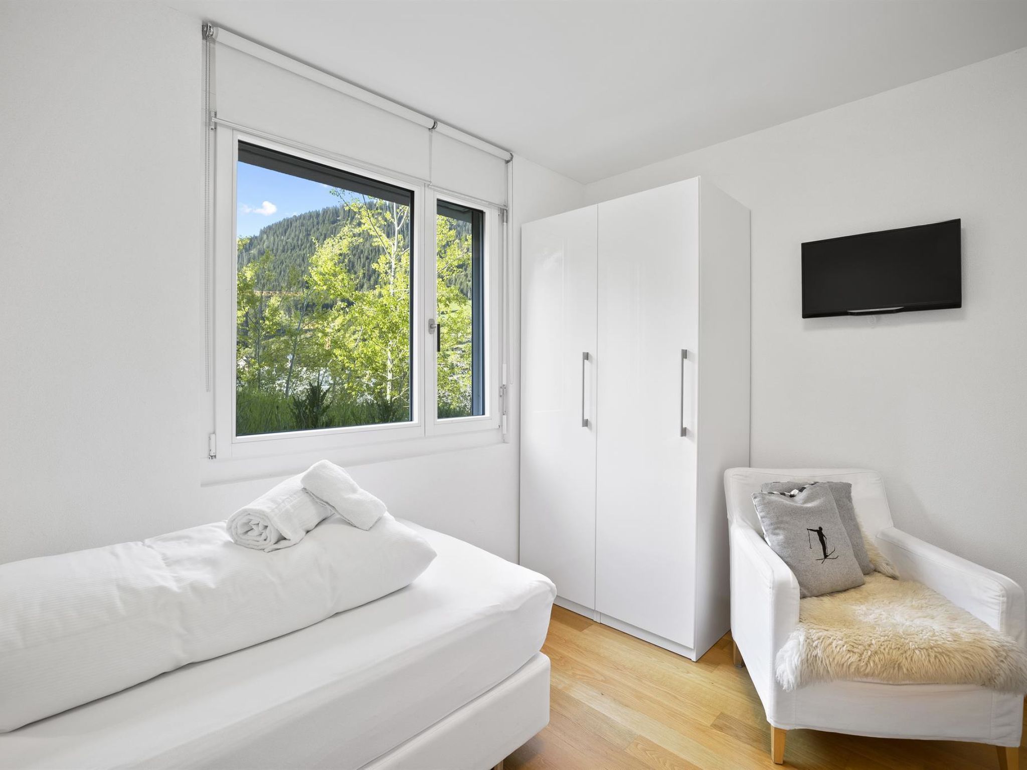 A bedroom with a bed, a chair, a wardrobe, and a television on the wall.