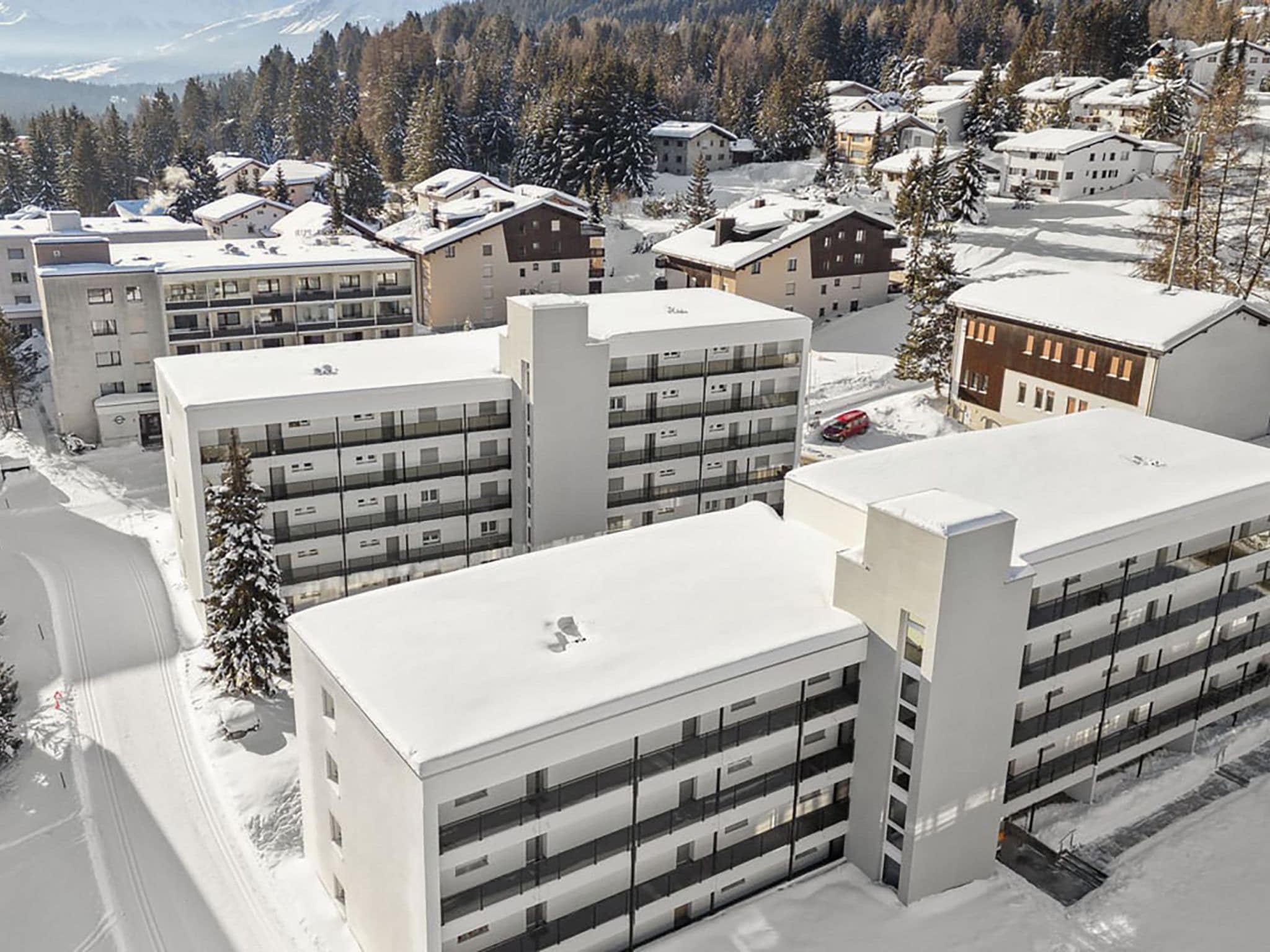 A modern multi-story building with a flat roof, likely a hotel or apartment complex, surrounded by snow-covered trees and a mountainous backdrop.