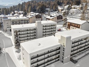 A modern multi-story building with a flat roof, likely a hotel or apartment complex, surrounded by snow-covered trees and a mountainous backdrop. - Nr.55 Haus Sotmirana Appartement/Fewo,   Nr.55 Haus Sotmirana by Interhome (Lenzerheide)