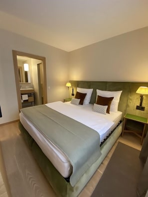 Ein Hotelzimmer mit Bett, zwei Nachttischlampen, einem Nachttisch und einem durchgegangenen Türrahmen einsehbaren Badezimmer.