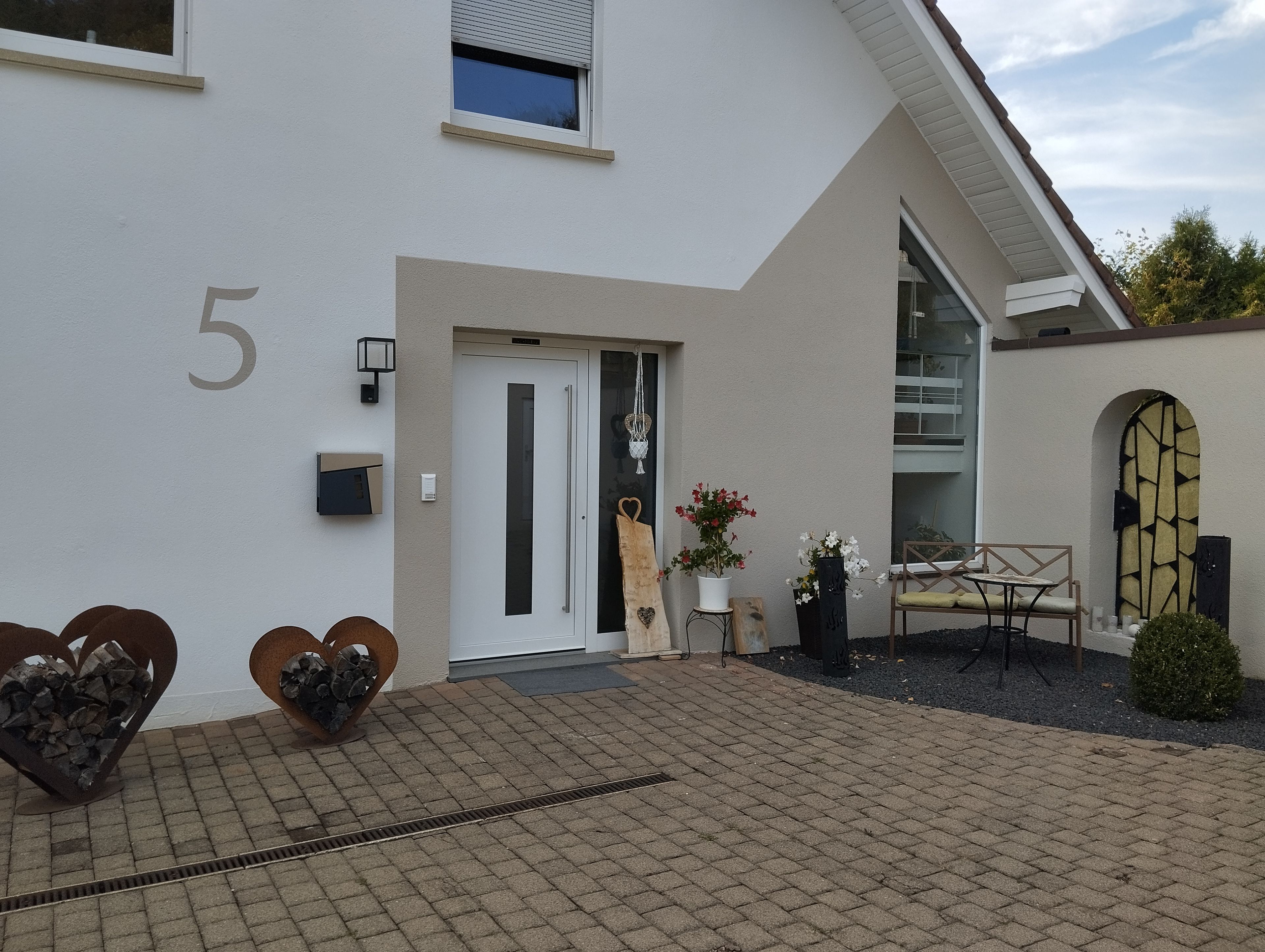 A modern building with a number '5' on the wall, a door, a small outdoor seating area with a table and chairs, and decorative heart-shaped planters.