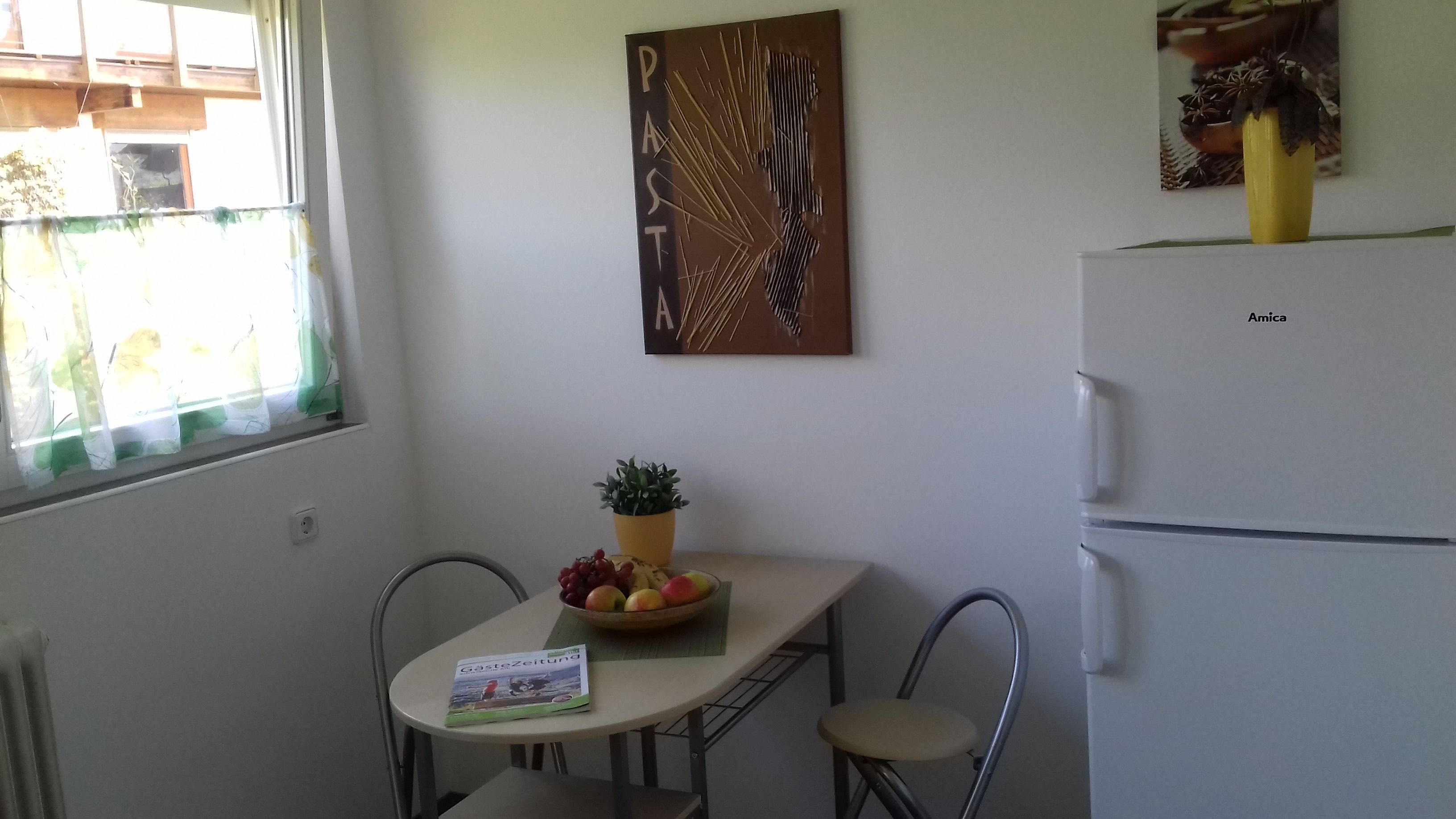 A small kitchen with a white refrigerator, a table with chairs, a potted plant, and a wall-mounted artwork.