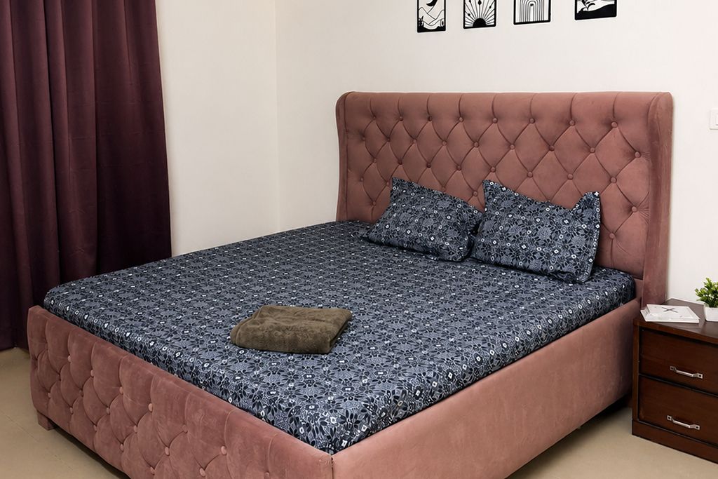 Een slaapkamer met een geborduurde hoofdbord, een bed met een gedessineerd sprei, twee kussens, een klein nachtkastje met een plant, en een donker gordijn.