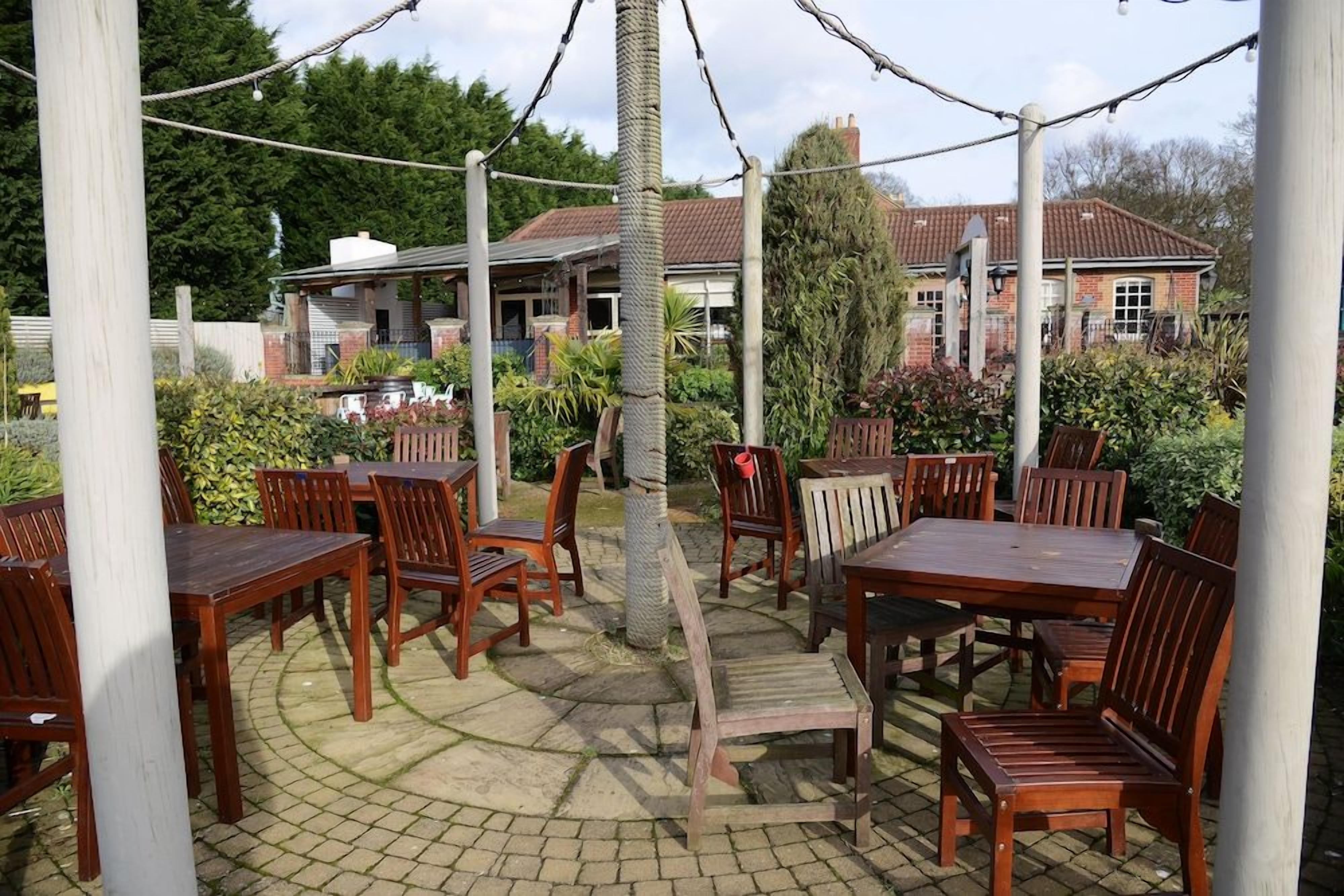 An outdoor patio area with wooden tables and chairs, surrounded by greenery and a building in the background.
