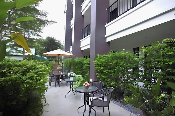 Un espace extérieur aménagé en terrasse, avec des tables et des chaises, entouré de verdure et bordé d’un bâtiment doté de balcons.