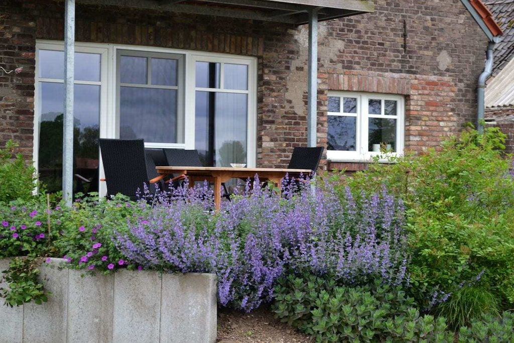 Edificio de ladrillo con un patio que incluye una mesa y sillas, rodeado por un jardín de flores moradas.