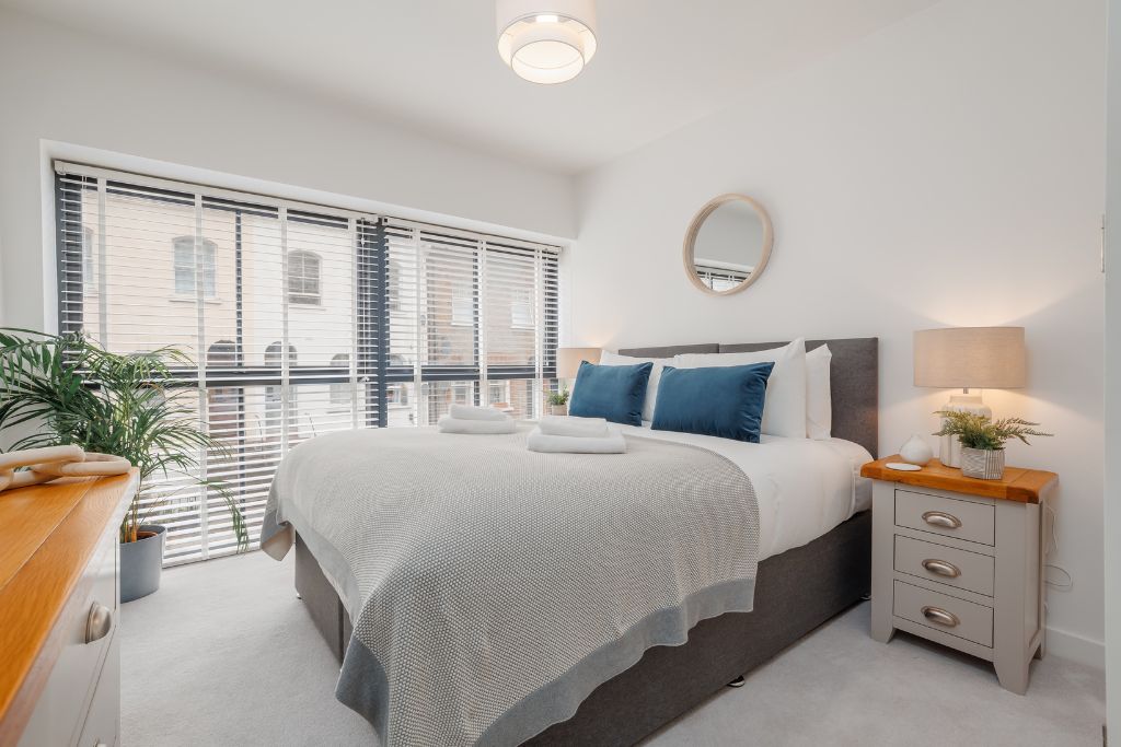 Ein Schlafzimmer mit Bett, Nachttisch, Lampe und Blick ins Freie durch große Fenster.