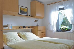 Ein Schlafzimmer mit zwei Betten, einer hölzernen Kommode, einem Schreibtisch mit Lampe, einem Regal mit Büchern und Dekorationsgegenständen sowie einem Fenster mit Vorhängen.