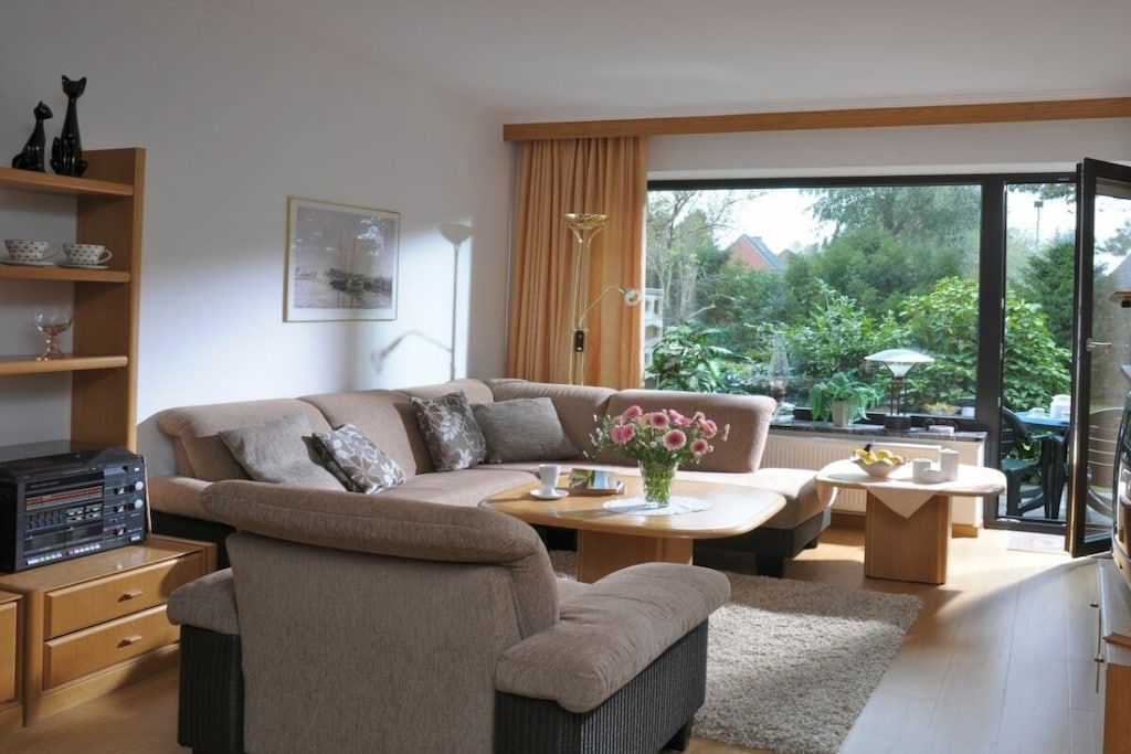 A modern living room with a sofa, coffee table, and a television. There is a bookshelf with decorative items and a window with a view of greenery.