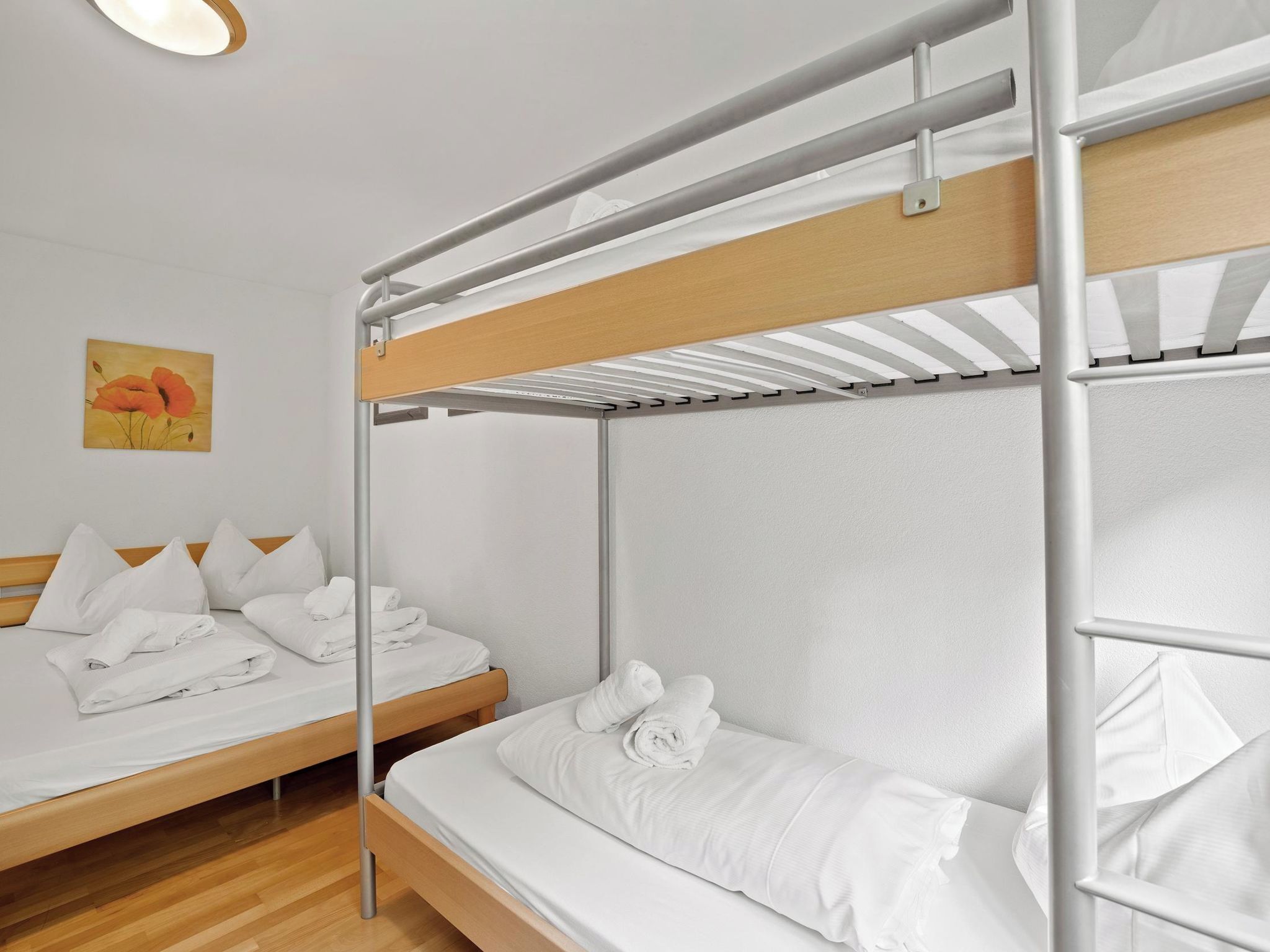 Un lit superposé avec une literie blanche et une tête de lit en bois, un lit simple avec une literie blanche et des oreillers, et un tableau représentant une fleur de coquelicot accroché au mur.