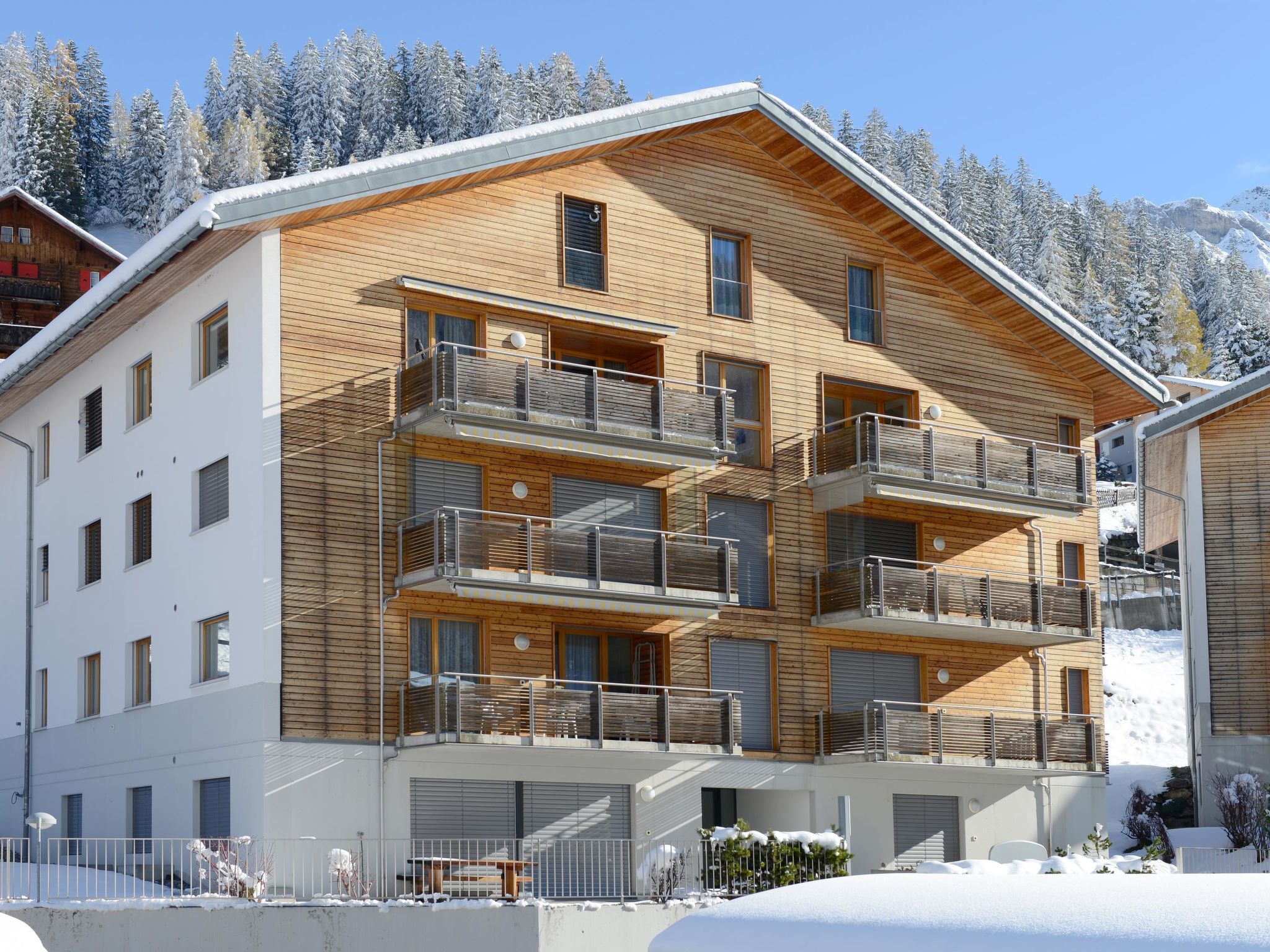 A multi-story wooden building with balconies, a covered entrance, and a view of snow-covered trees.