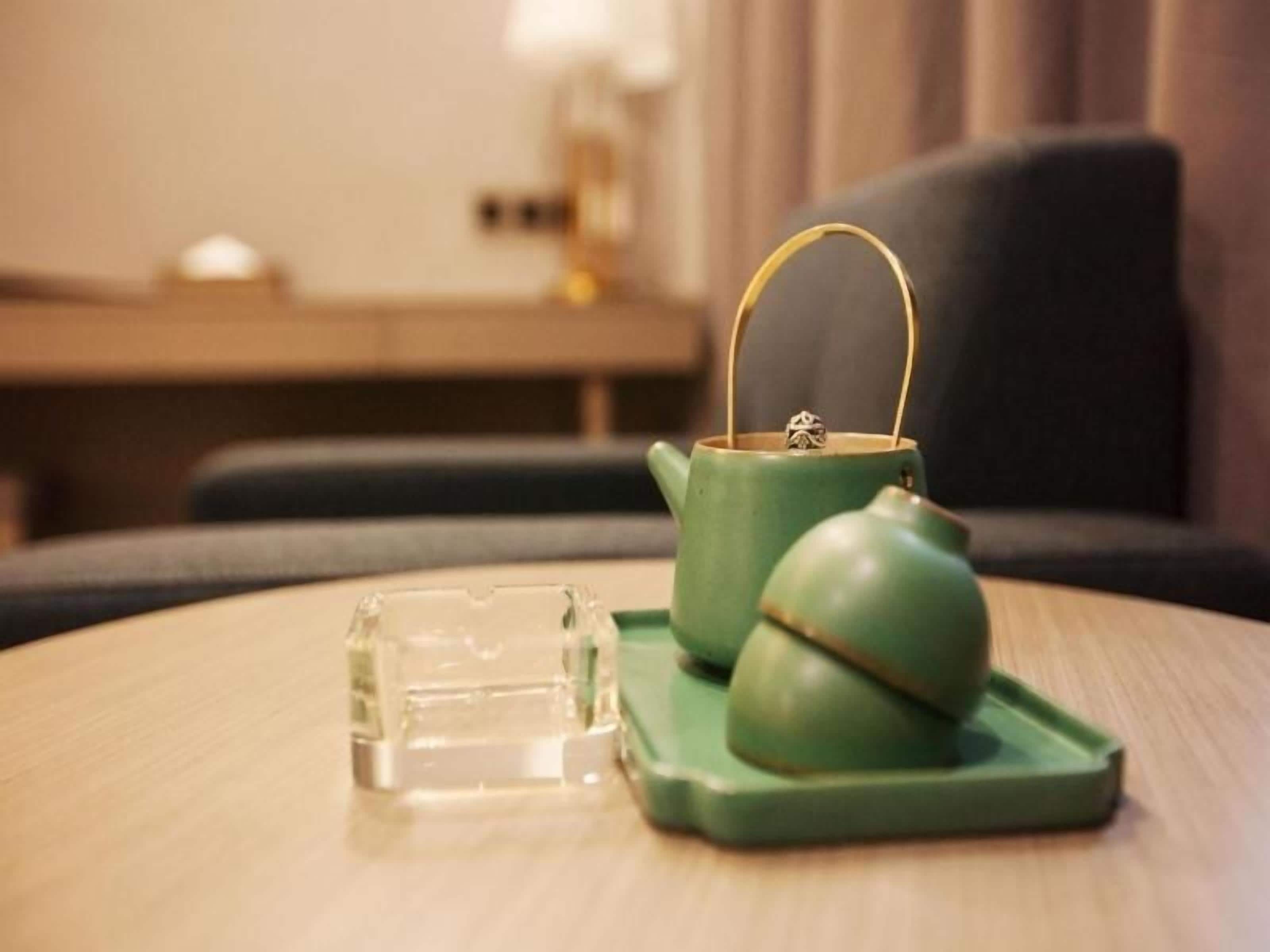 Een groene theepot en kopje op een tafel, met een glazen vaas ernaast.