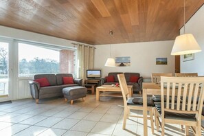 Ein modernes Wohnzimmer mit Holzboden, einem Fernseher, einem Sofa, einem Couchtisch und einem Essbereich mit Holzstühlen.