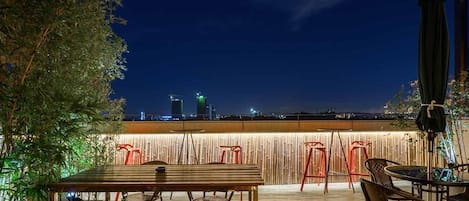 Un patio al aire libre con una mesa y sillas de madera, una valla de bambú y un horizonte urbano de fondo.