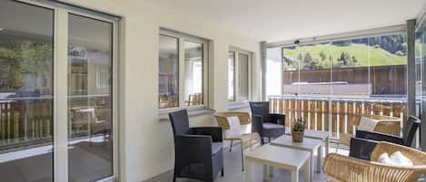 A modern outdoor patio with wicker furniture, a glass door leading to a balcony, and a view of a hilly landscape.