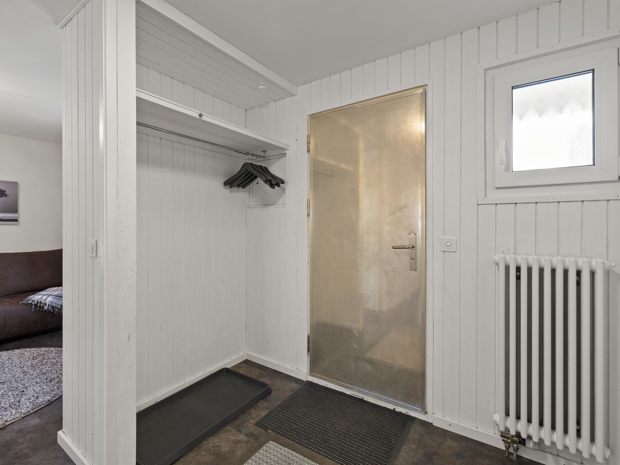 Una habitación con una puerta blanca, un radiador, una ventana y un perchero con ropa.