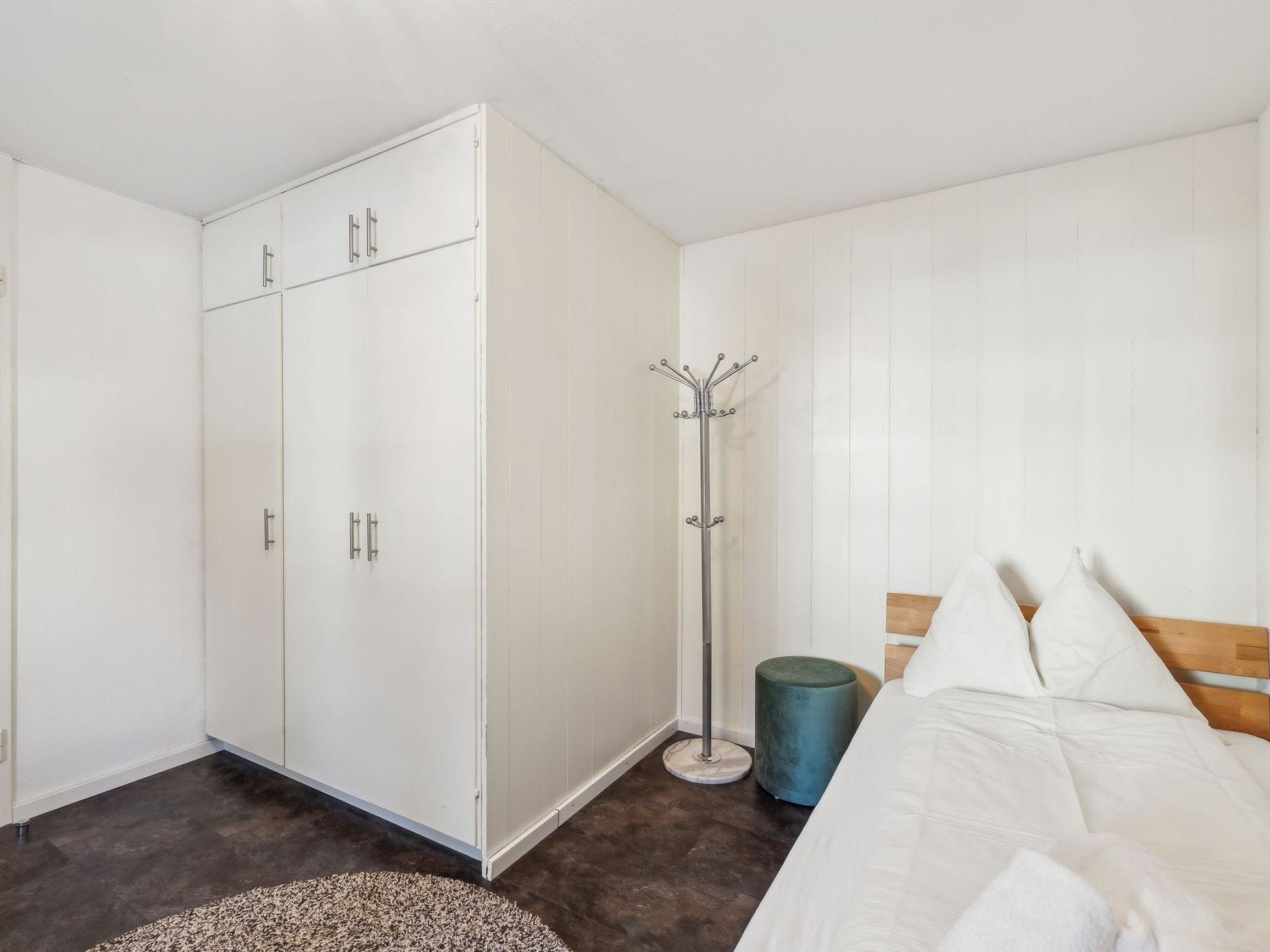 A bedroom with a bed, a wardrobe, a coat rack, and a round blue stool.