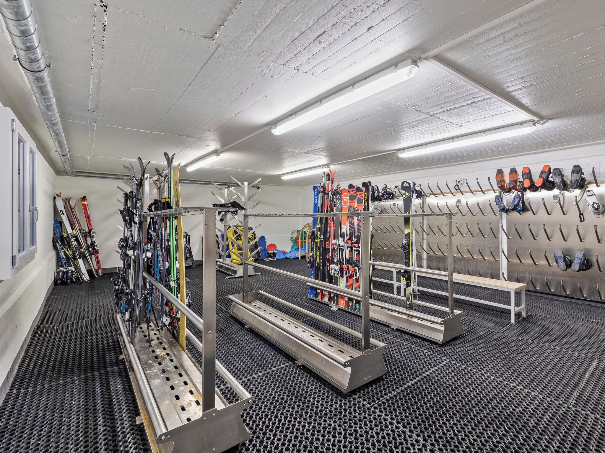 Een skiopslag met rekken voor ski's en snowboards, een bankje en een ventilatiesysteem.
