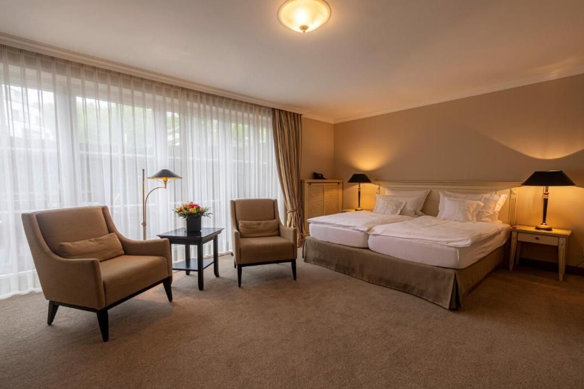 A hotel room with a double bed, two armchairs, a small table with a vase, and two bedside lamps.