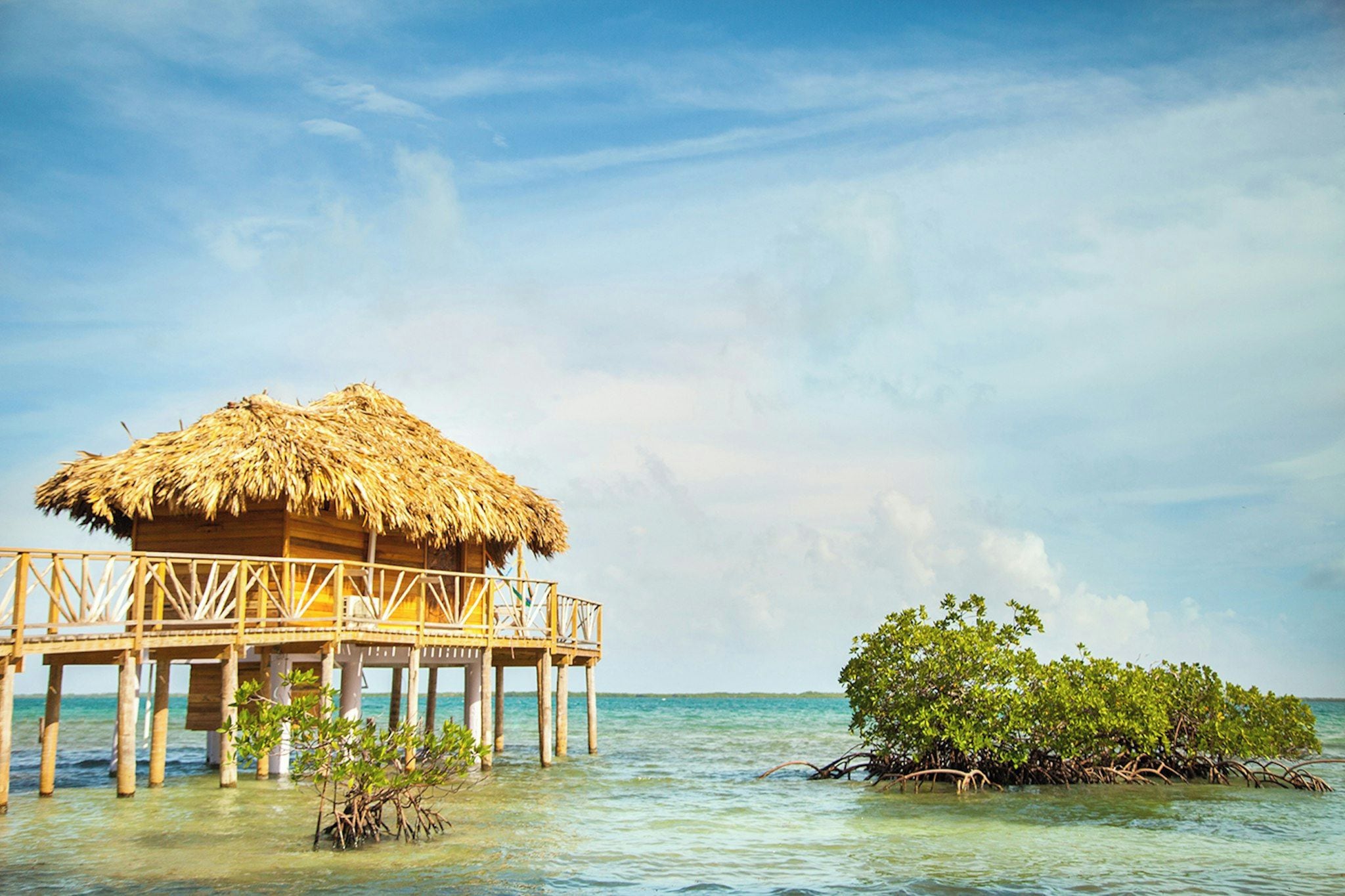 A wooden overwater bungalow with a thatched roof, surrounded by clear blue water and a mangrove island.