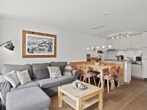 Uma sala de estar moderna com um sofá cinza, uma mesa de centro de madeira e uma área de cozinha com uma mesa de jantar e cadeiras.